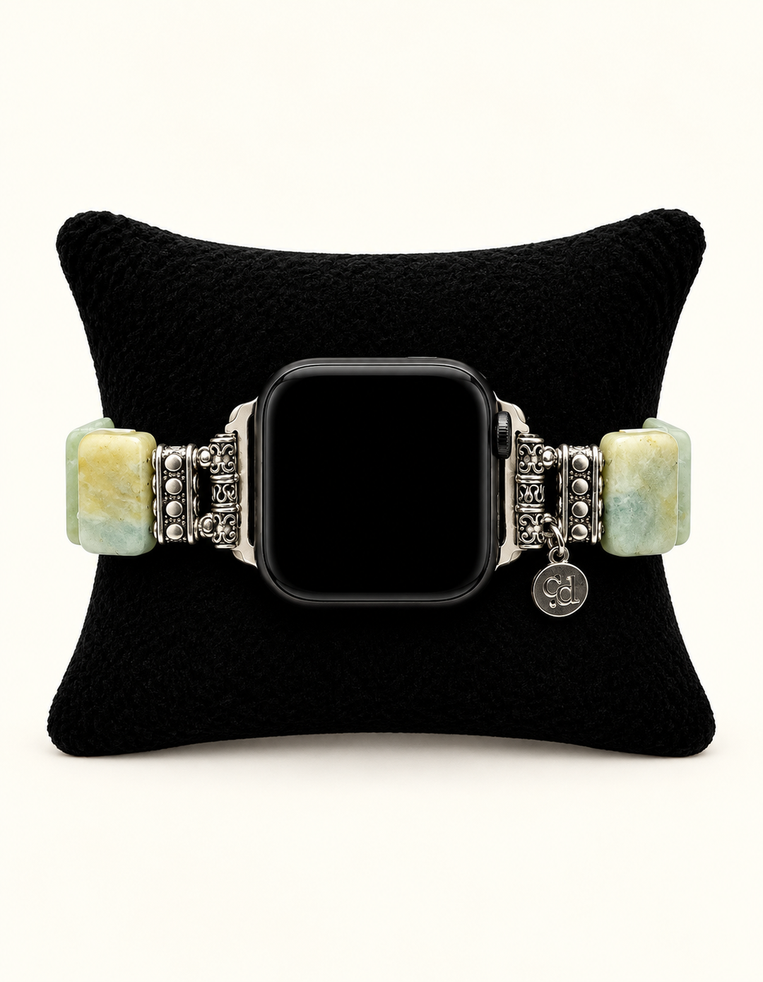 A silver-toned metal bracelet watch with rectangular light green and yellow stone beads, displayed on a black velvet cushion. The watch face is black and blank.