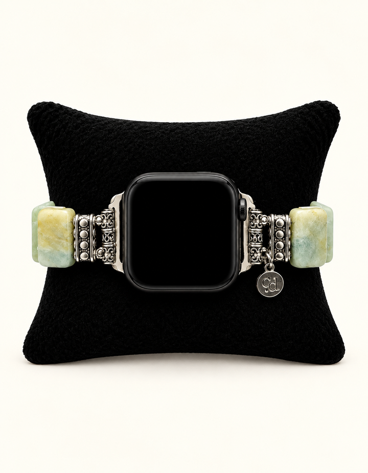 A silver-toned metal bracelet watch with rectangular light green and yellow stone beads, displayed on a black velvet cushion. The watch face is black and blank.