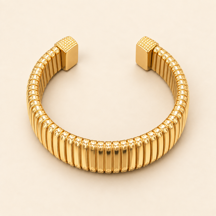 Open gold Omega cuff bracelet with vertical ribbed detailing and textured square end caps flat-laid on a cream background.