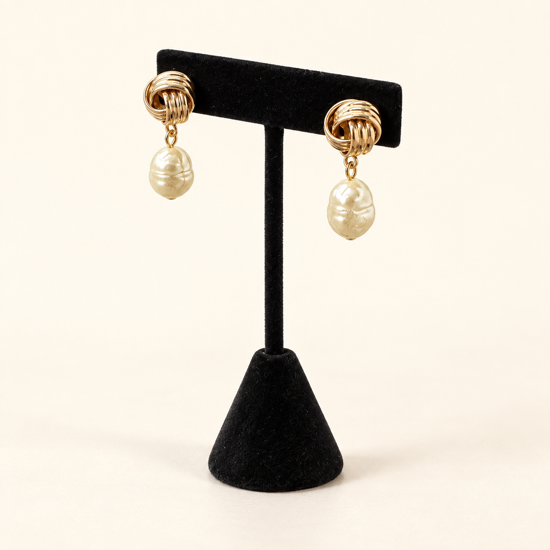 Angled view of textured gold knot earrings with organic ivory baroque pearl drops on a black velvet stand.
