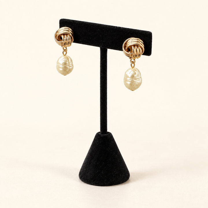 Angled view of textured gold knot earrings with organic ivory baroque pearl drops on a black velvet stand.