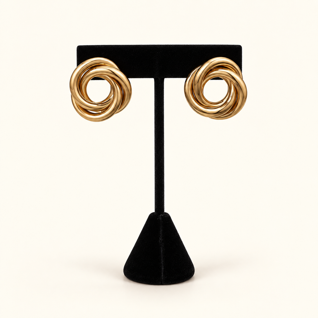 Front view of oversized gold-tone Love Knot Metal Earrings with smooth interwoven loops on a black velvet stand against a cream background.