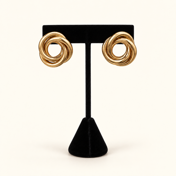 Front view of oversized gold-tone Love Knot Metal Earrings with smooth interwoven loops on a black velvet stand against a cream background.
