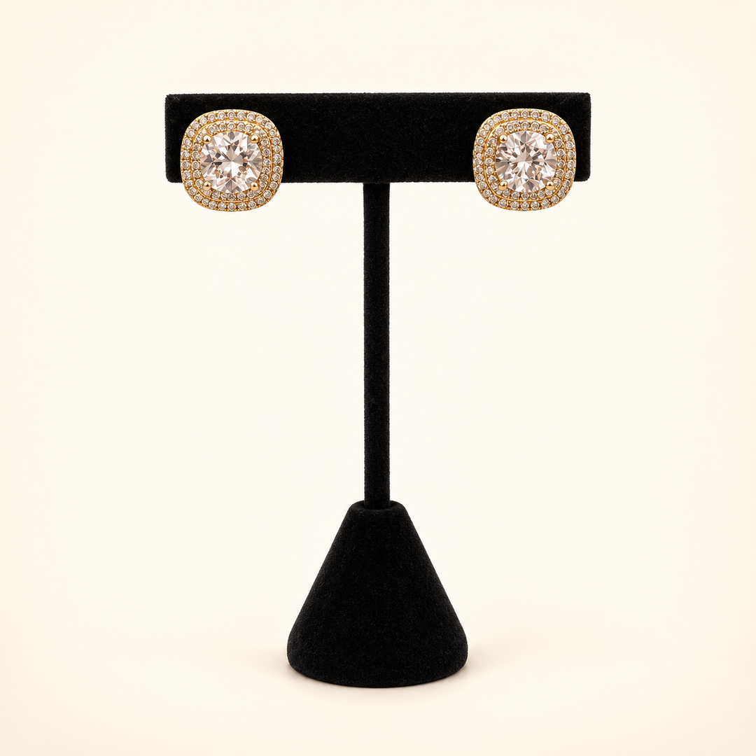 Gold square CZ halo stud earrings with large clear center stones displayed on a black velvet stand against a cream background.