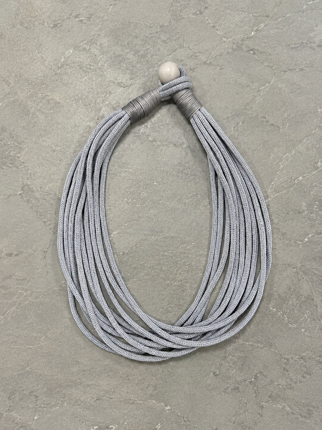 Overhead flat lay of the light gray multi-strand cord necklace highlighting the wrapped knot closure and layered woven cord design on a gray stone surface.