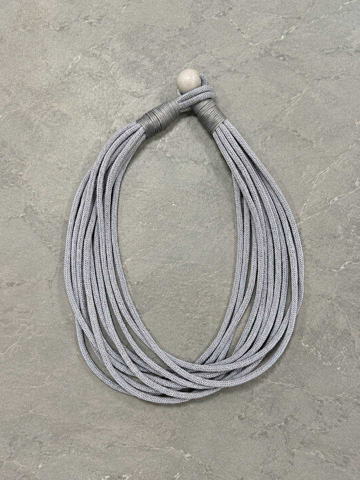 Overhead flat lay of the light gray multi-strand cord necklace highlighting the wrapped knot closure and layered woven cord design on a gray stone surface.