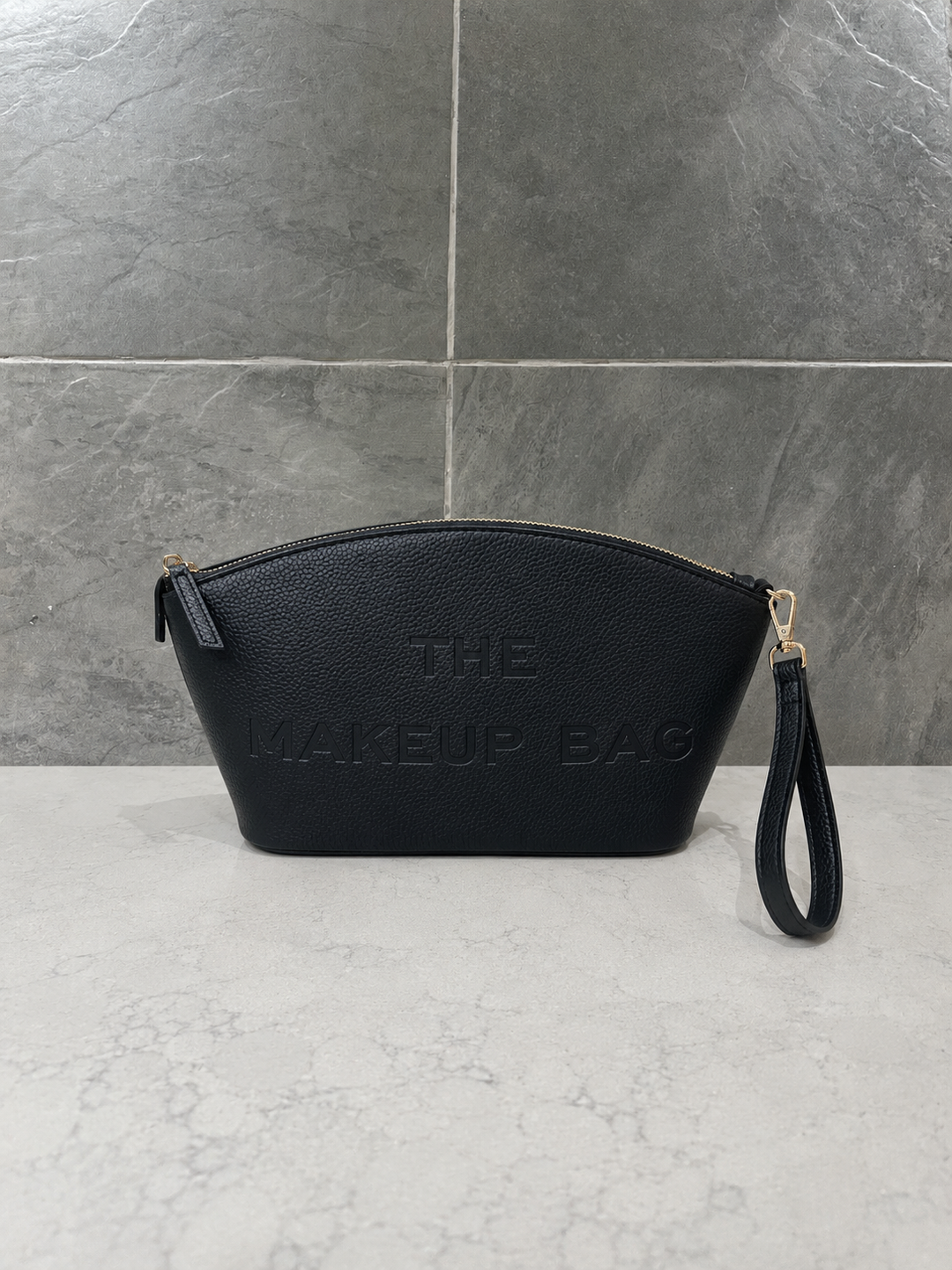 Black pebbled faux leather makeup bag with embossed “THE MAKEUP BAG” lettering, gold-tone zipper and detachable wristlet strap, displayed on a light marble countertop with a modern gray stone tile backdrop.