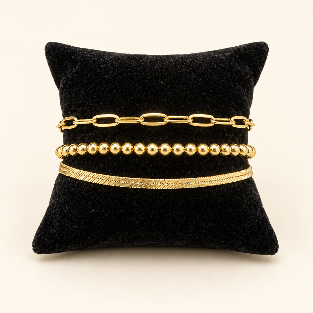Gold three-row bracelet featuring paperclip links, polished beads, and a smooth snake chain displayed on a black velvet pillow against a cream background.