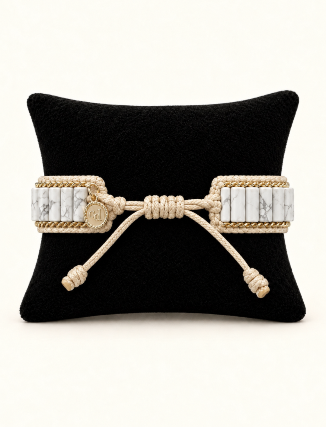 A wide woven rope bracelet with rectangular white marble inlays and a central knotted bow detail, displayed on a black cushion against a light beige background.