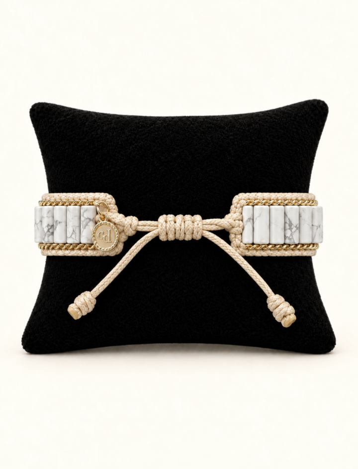 A wide woven rope bracelet with rectangular white marble inlays and a central knotted bow detail, displayed on a black cushion against a light beige background.
