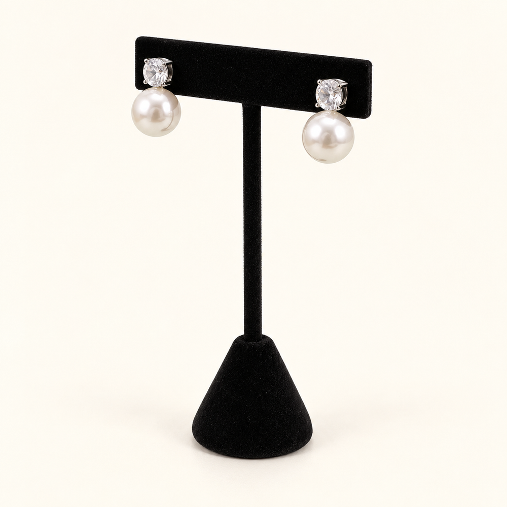 Angled view of round crystal and ivory pearl drop post earrings on a black velvet stand against a cream background.