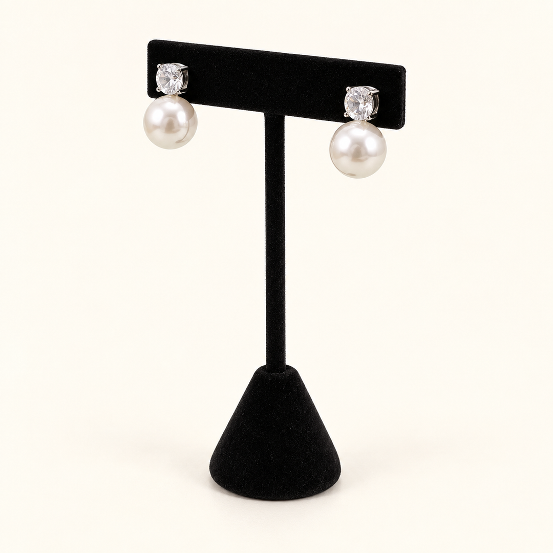 Angled view of round crystal and ivory pearl drop post earrings on a black velvet stand against a cream background.