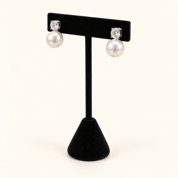 Angled view of round crystal and ivory pearl drop post earrings on a black velvet stand against a cream background.