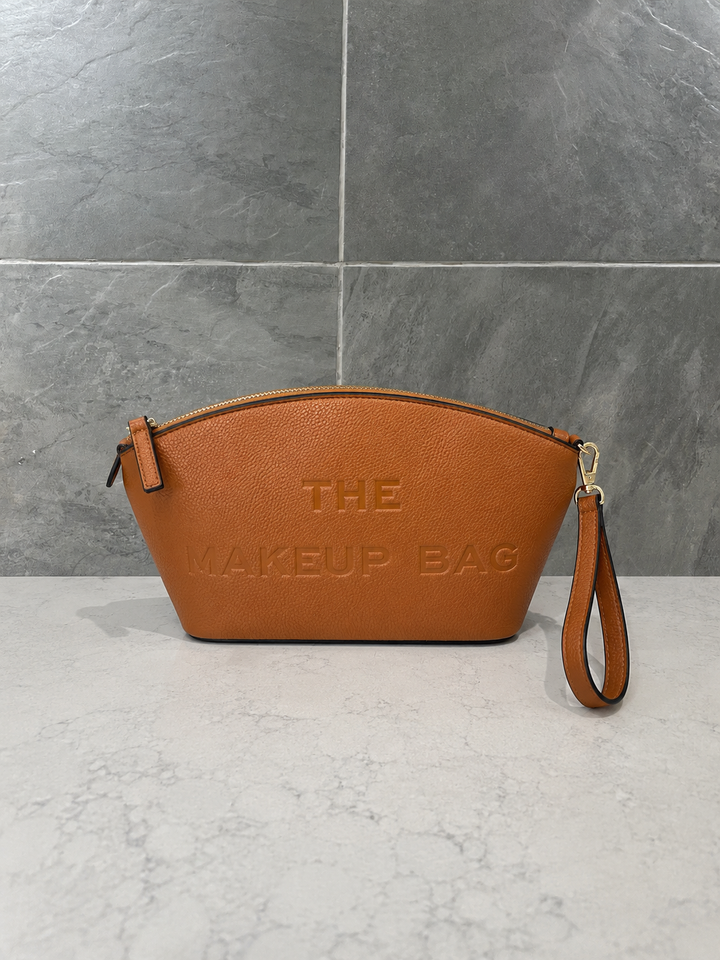Camel pebbled faux leather makeup bag with embossed “THE MAKEUP BAG” lettering, gold-tone zipper and detachable wristlet strap, photographed on a light marble countertop with a textured gray stone tile wall.