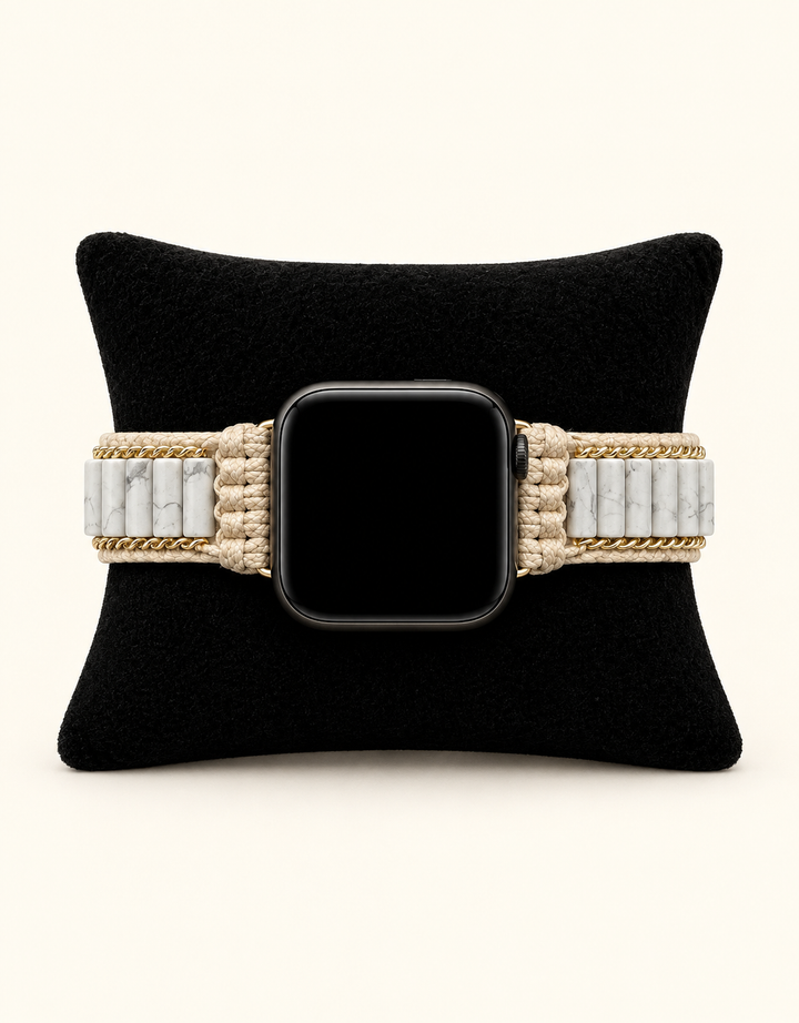 A woven watch band in beige and white with gold-toned chain accents, shown on a black watch with a black screen, displayed on a black velvet cushion against a light cream background.