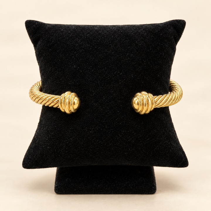 Gold textured metal cuff bracelet with twisted cable detailing and ridged end caps displayed on a black velvet pillow stand.
