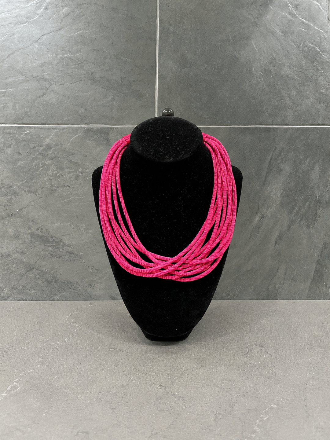 Hot pink multi-strand cord statement necklace showcased on a black velvet jewelry bust against a gray stone background.