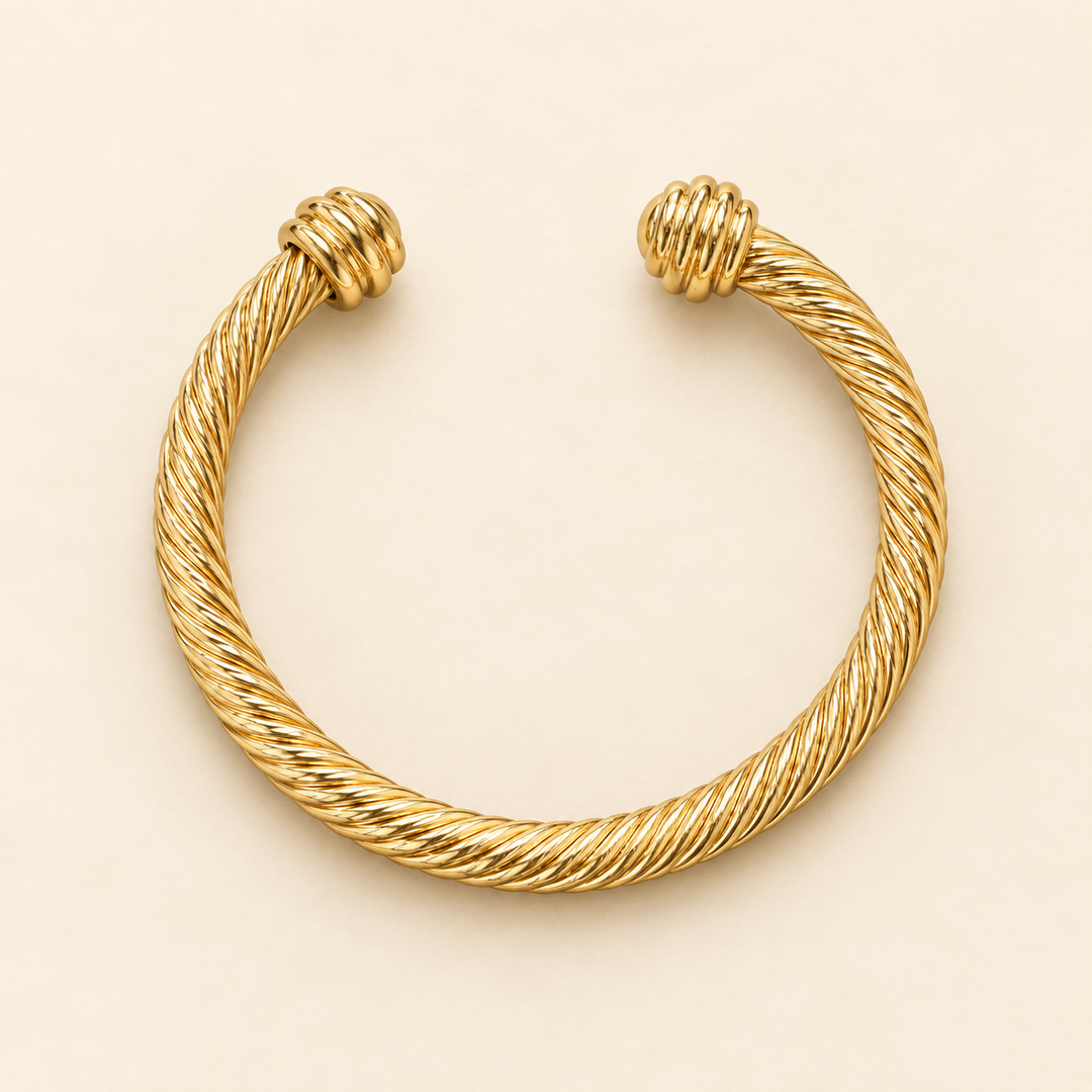 Open gold textured metal cuff bracelet with twisted rope detailing and rounded ridged end caps flat-laid on a cream background.
