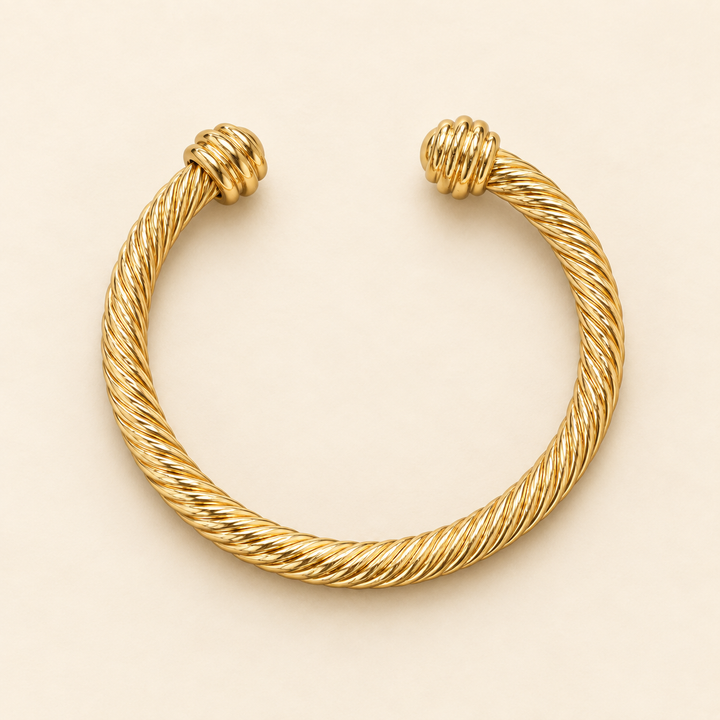 Open gold textured metal cuff bracelet with twisted rope detailing and rounded ridged end caps flat-laid on a cream background.