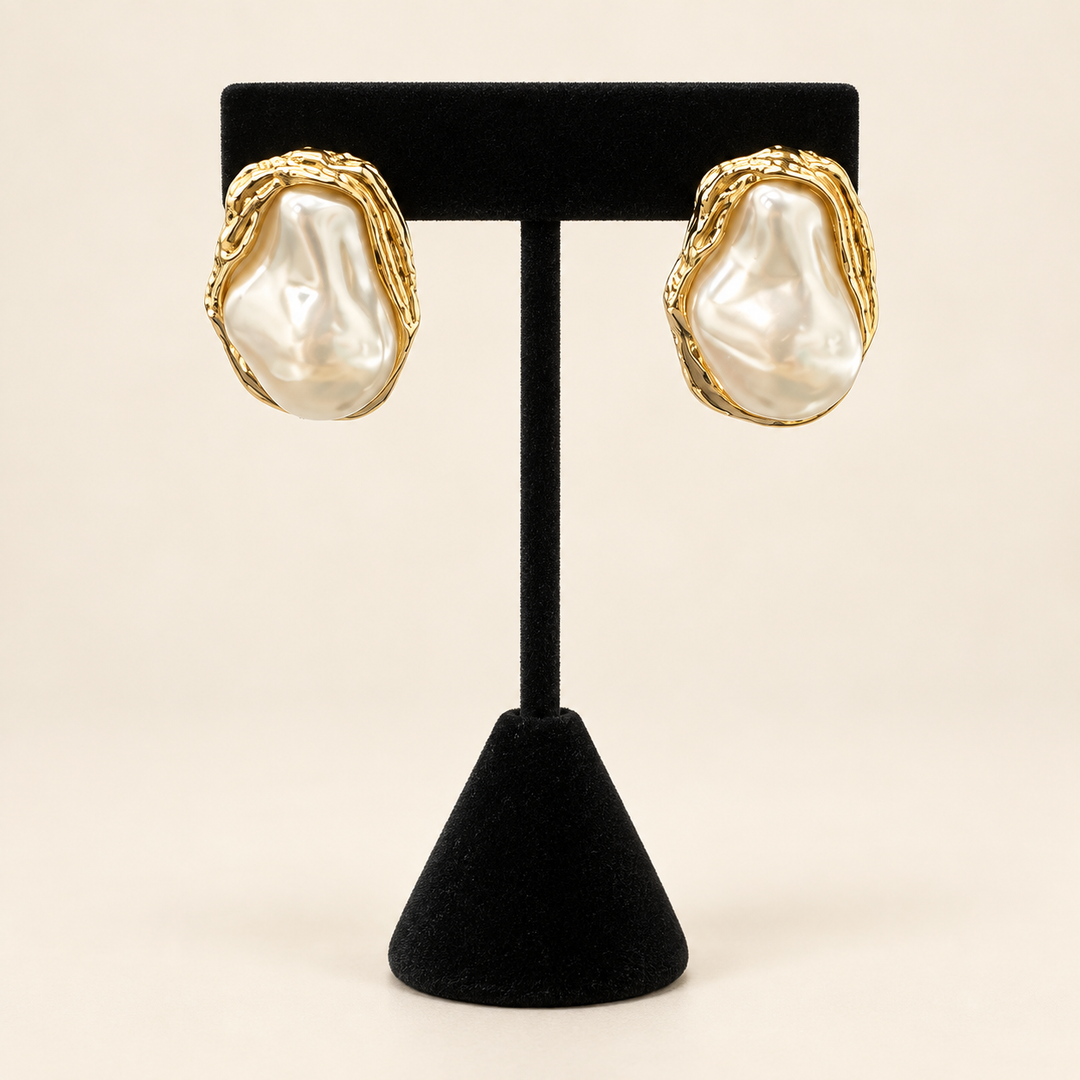 Front view of ivory baroque pearl stud earrings with sculptural textured gold-tone frames displayed on a black velvet stand.
