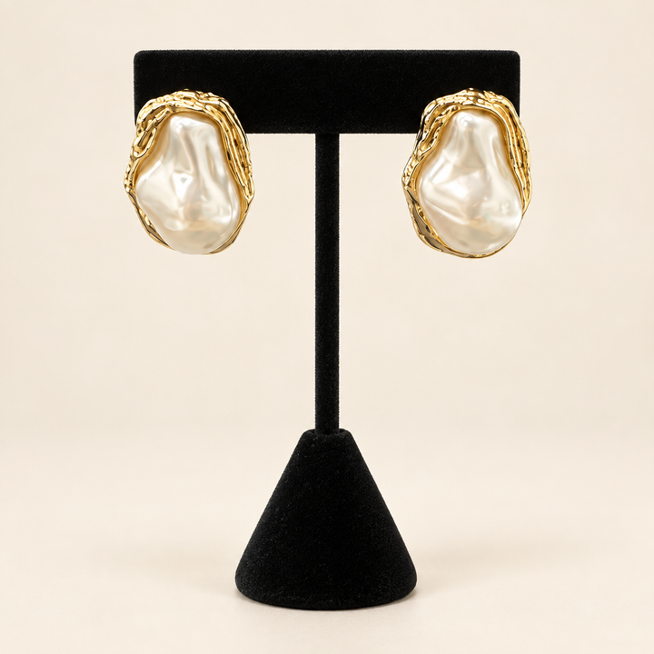 Front view of ivory baroque pearl stud earrings with sculptural textured gold-tone frames displayed on a black velvet stand.
