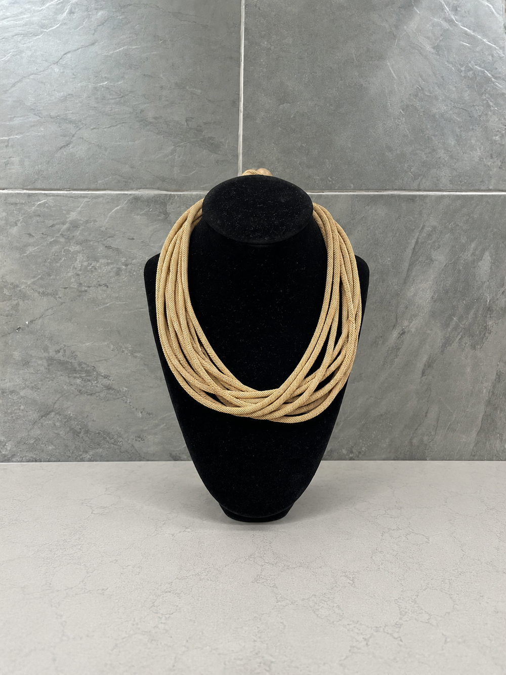 Light tan multi-strand woven cord statement necklace on a black velvet display with a gray textured stone background.