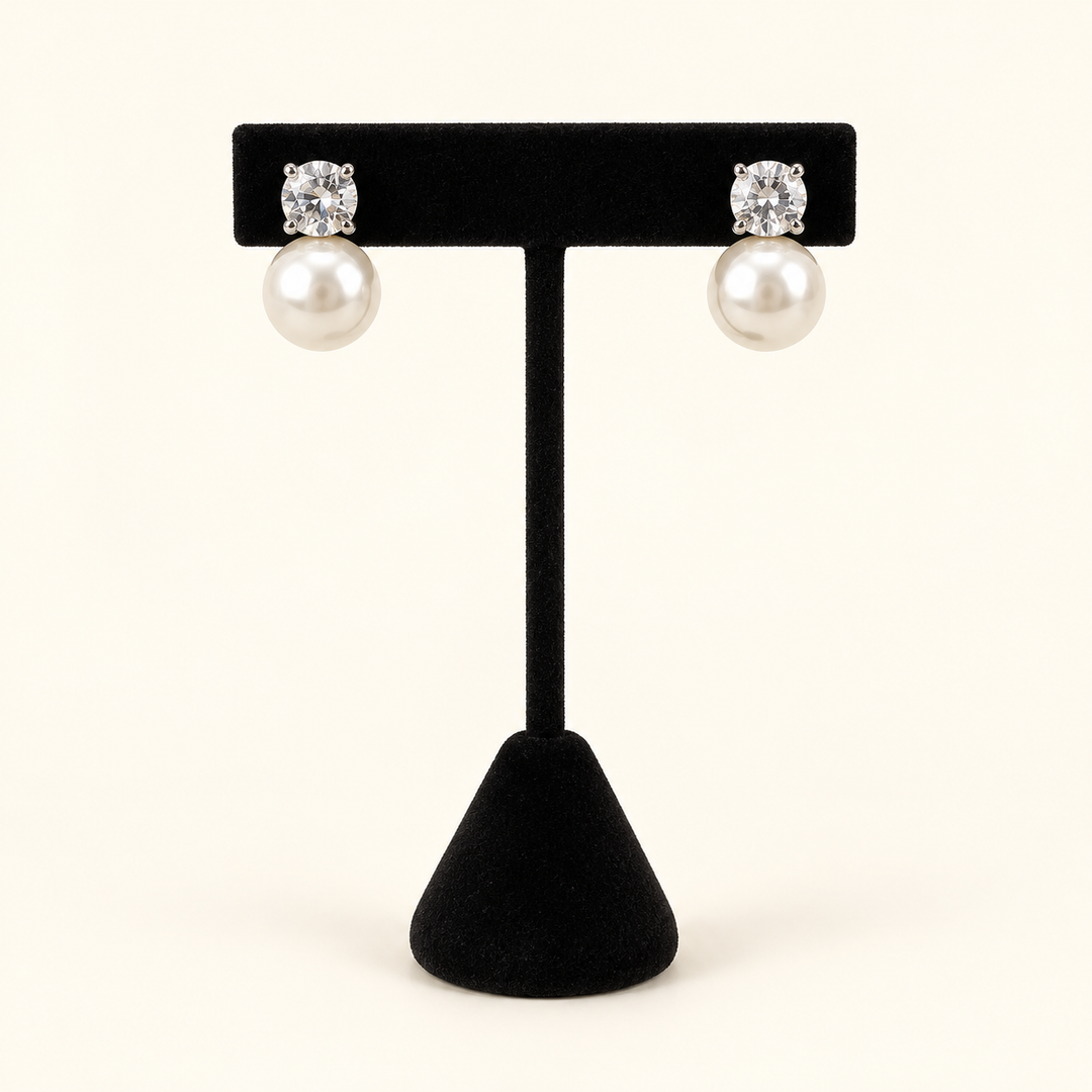 Front view of round clear crystal stud earrings with large ivory pearl drops on a black velvet stand against a cream background.
