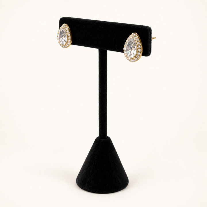 Angled view of gold teardrop halo stud earrings with clear faceted stones displayed on a black velvet stand.
