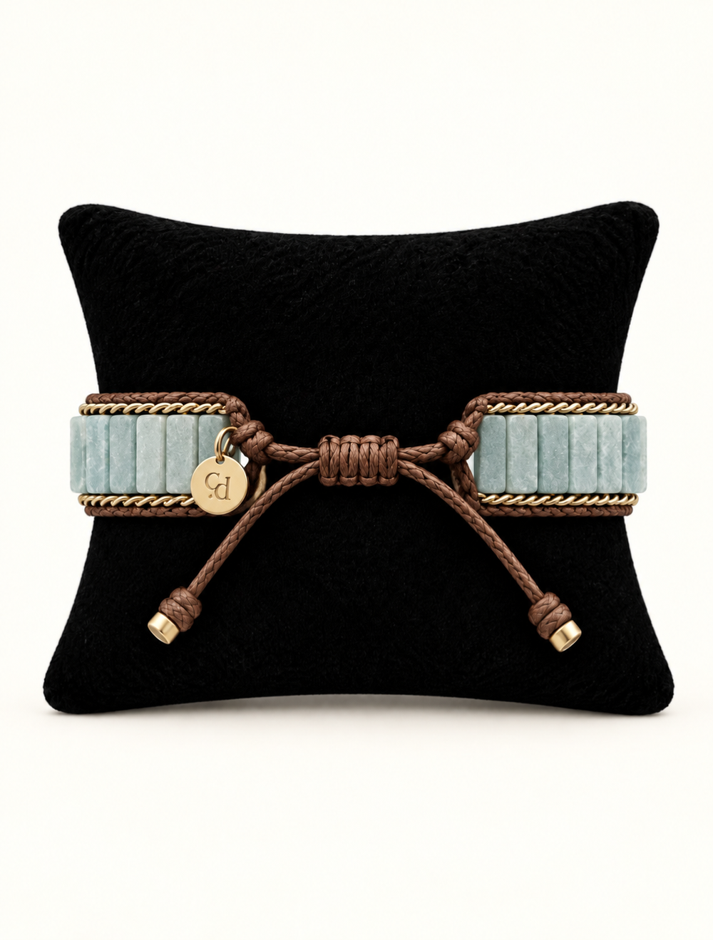 A wide bracelet with light blue ribbed bands, brown braided rope detailing, and a small round gold charm engraved with initials, displayed on a black pillow against a white background.