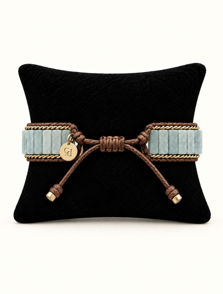 A wide bracelet with light blue ribbed bands, brown braided rope detailing, and a small round gold charm engraved with initials, displayed on a black pillow against a white background.