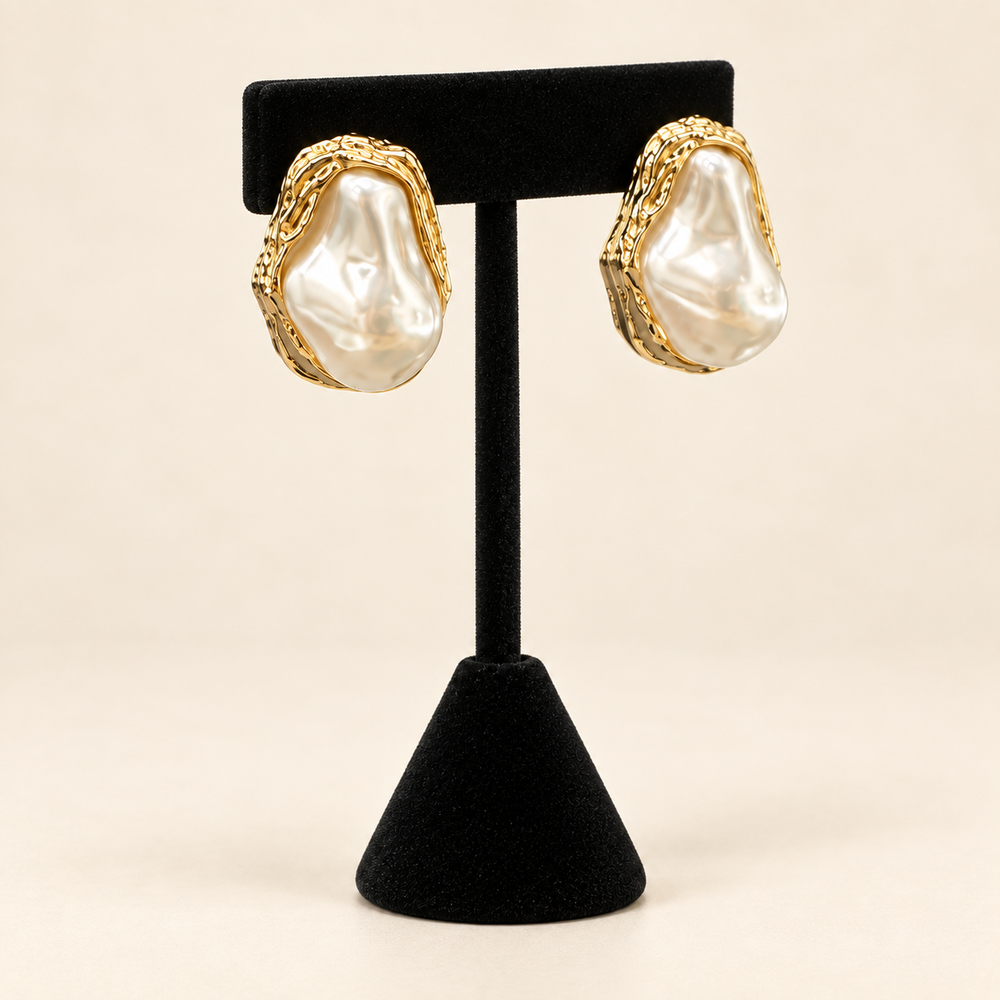 Angled view of framed ivory baroque pearl earrings showing their irregular shape and textured gold-tone edges on a black velvet stand.