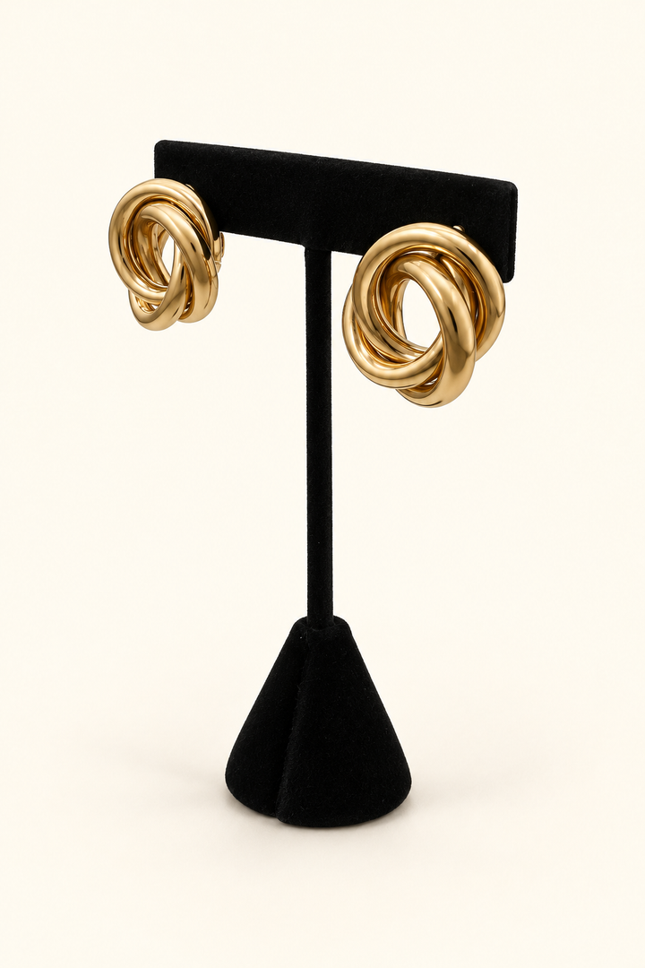 Angled view of oversized gold-tone Love Knot Metal Earrings showing the sculptural interwoven loop design on a black velvet stand against a cream background.