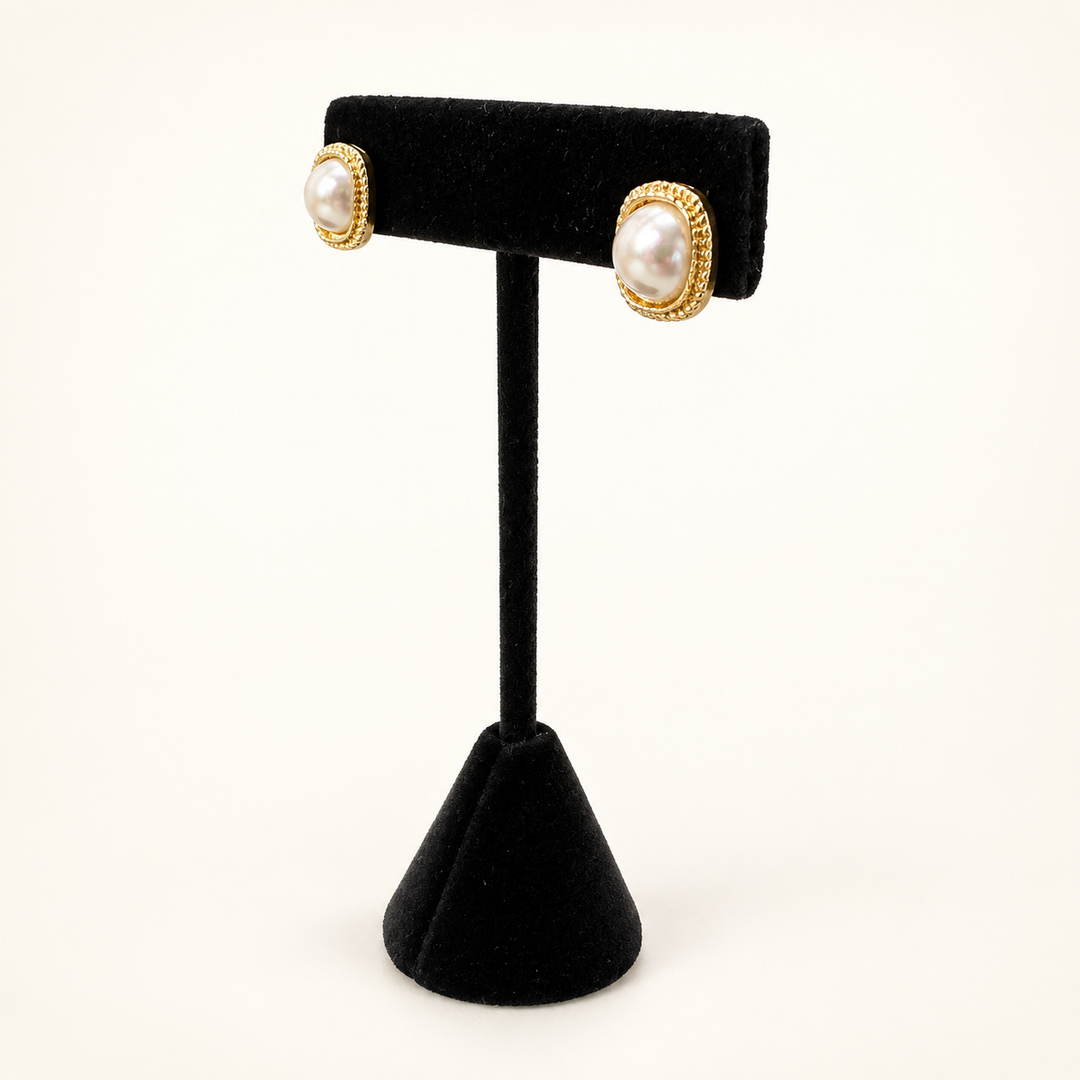 Angled view of gold square pearl stud earrings showing the domed ivory centers and textured frames on a black velvet stand.