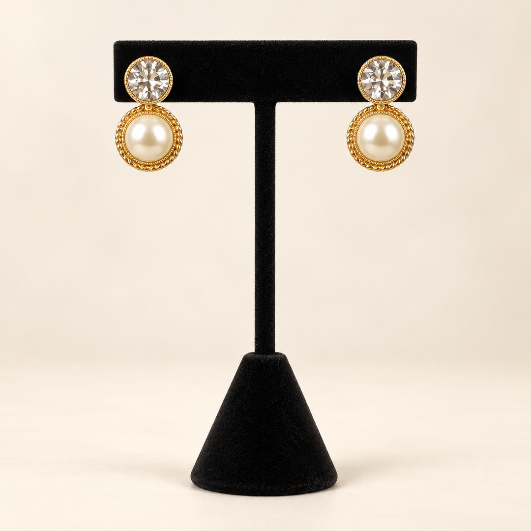 Front view of gold-tone round pearl textured drop earrings with clear crystal studs displayed on a black velvet stand.