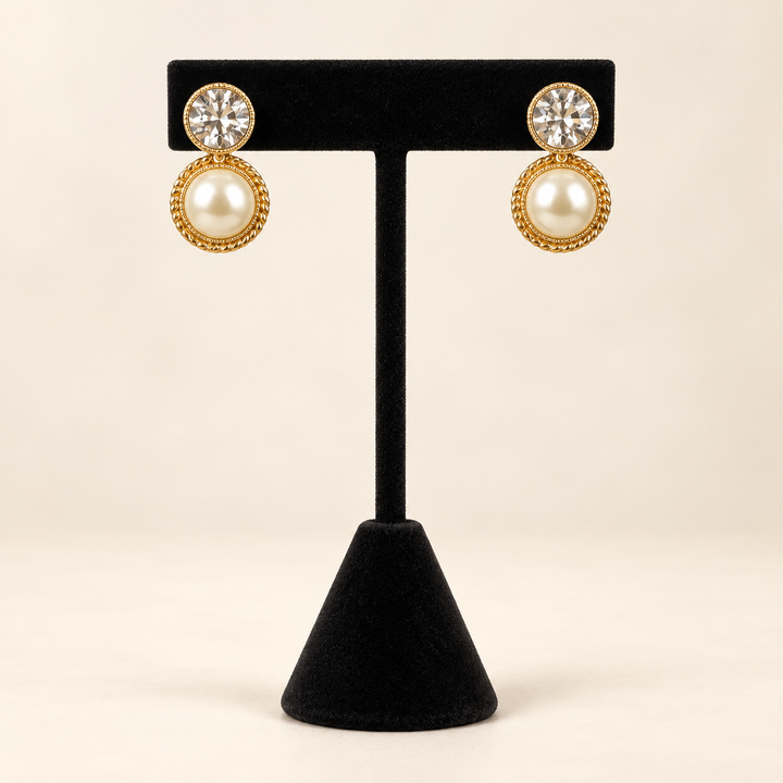 Front view of gold-tone round pearl textured drop earrings with clear crystal studs displayed on a black velvet stand.