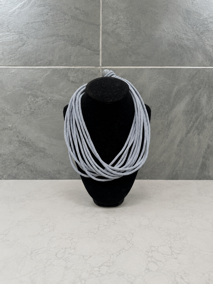 Light gray multi-strand cord statement necklace displayed on a black velvet jewelry bust against a modern gray stone backdrop.