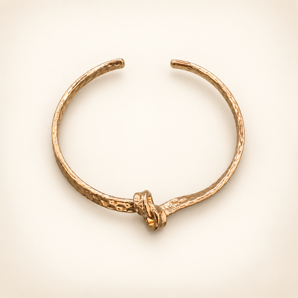 Hammered gold open cuff bracelet with a knotted center detail, displayed as a flat lay on a cream background.