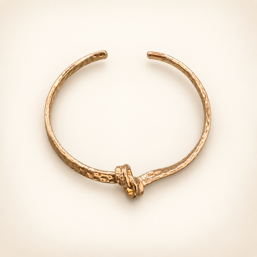 Hammered gold open cuff bracelet with a knotted center detail, displayed as a flat lay on a cream background.
