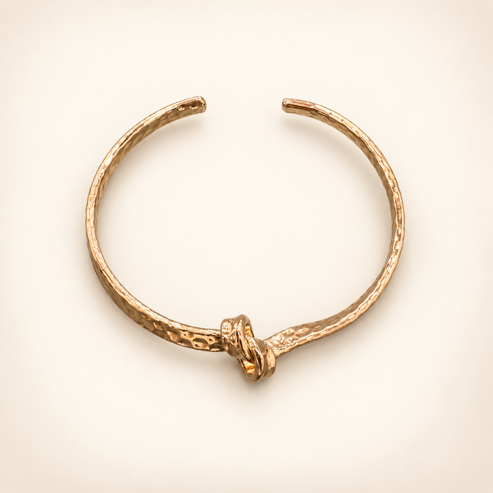 Hammered gold open cuff bracelet with a knotted center detail, displayed as a flat lay on a cream background.