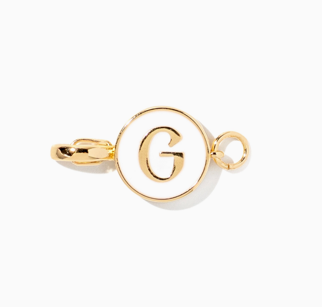 A gold initial charm featuring a circular bezel-set white enamel background with a polished gold letter &