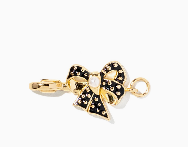 A gold bow-shaped pendant charm with black enamel sections, small rhinestone-like studs, and a central white pearl, shown on a plain white background.