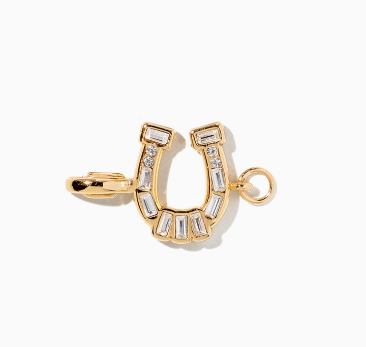 A gold horseshoe-shaped charm or pendant embellished with clear baguette and round crystals, shown on a clean white background.