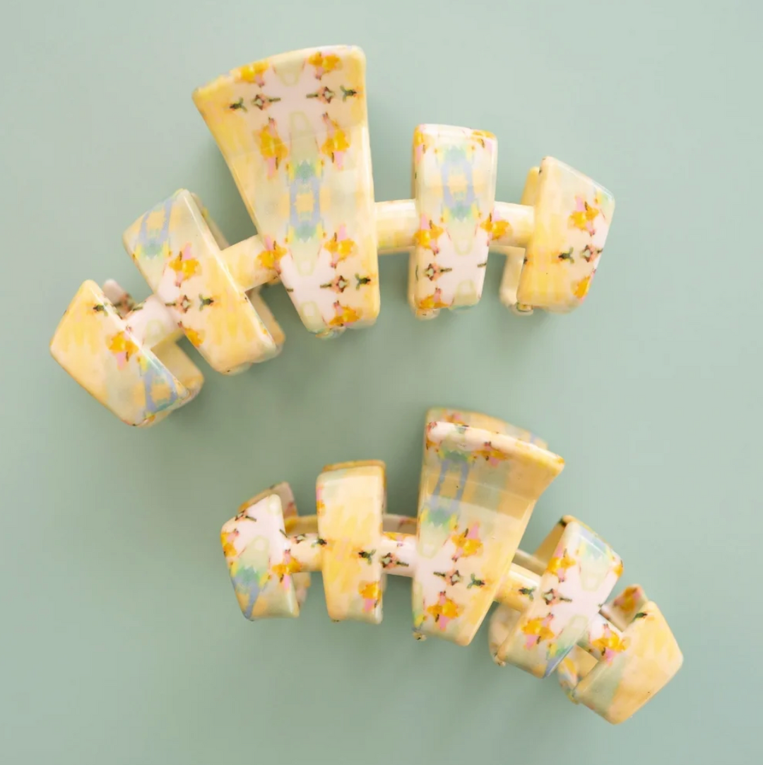 Two pale yellow hair claw clips with a delicate floral pattern, displayed on a solid sage green background.
