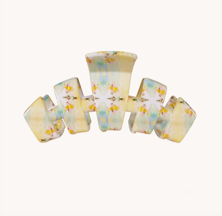 A curved hair claw clip with a glossy finish, decorated with a delicate pastel floral pattern in soft yellow, blue, and green on a white base, set against a plain off-white background.