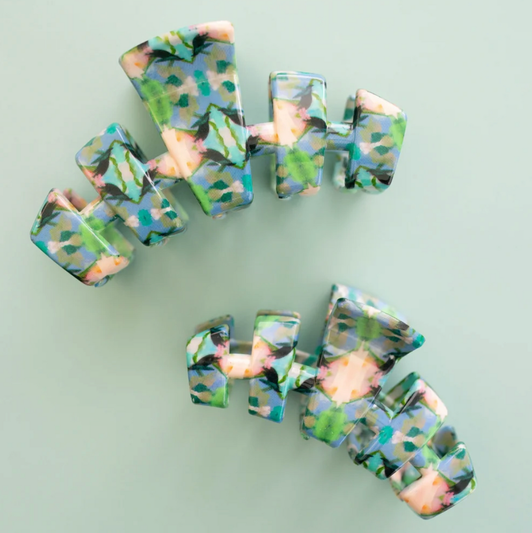 Two chunky resin claw hair clips with a marbled pattern of green, blue, black, and peach, displayed on a solid pale sage green background.