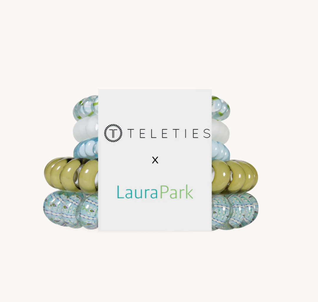 A set of four beaded stretch bracelets by Teleties x Laura Park, featuring a mix of olive green, light blue, and white beads, wrapped in a white paper band with the brand logos.
