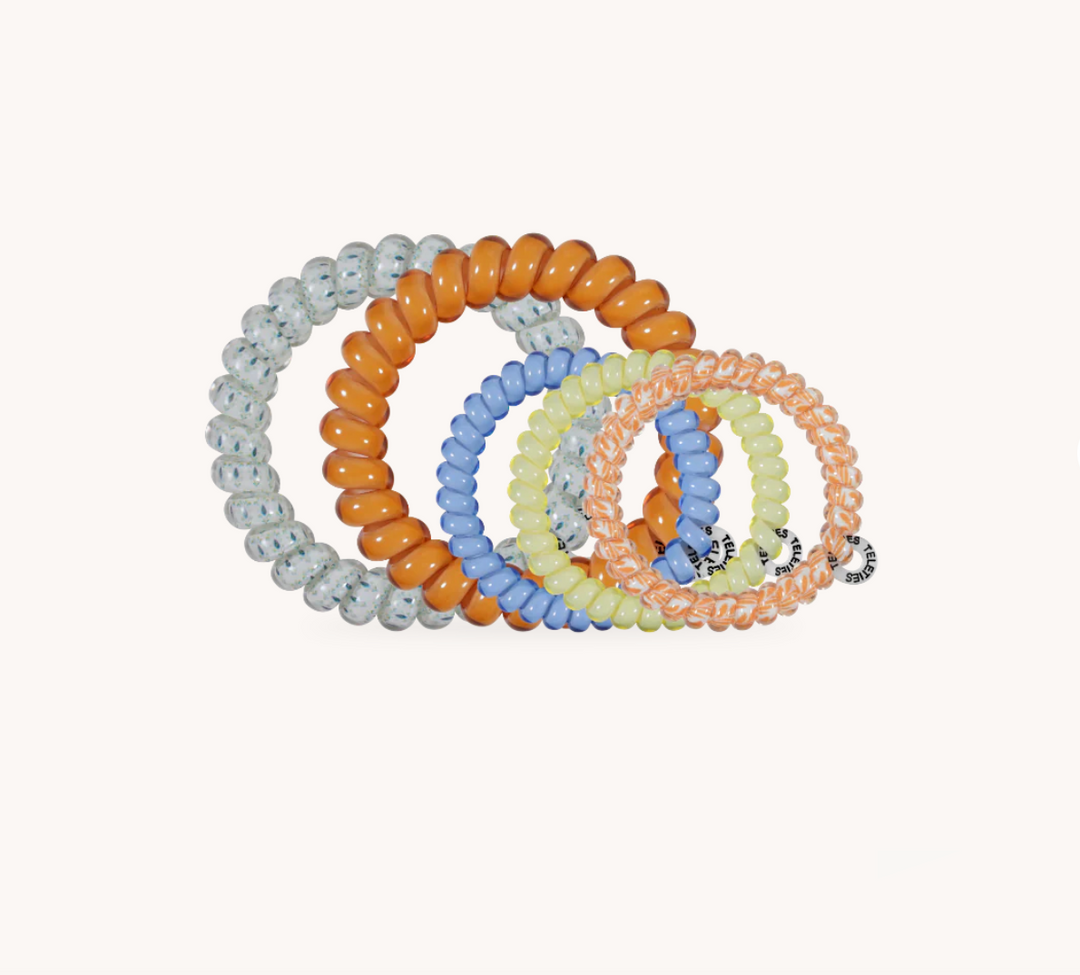 A set of five coiled elastic hair ties in assorted pastel and neutral colors, including light gray, orange, sky blue, pale yellow, and peach, arranged on a solid off-white background.
