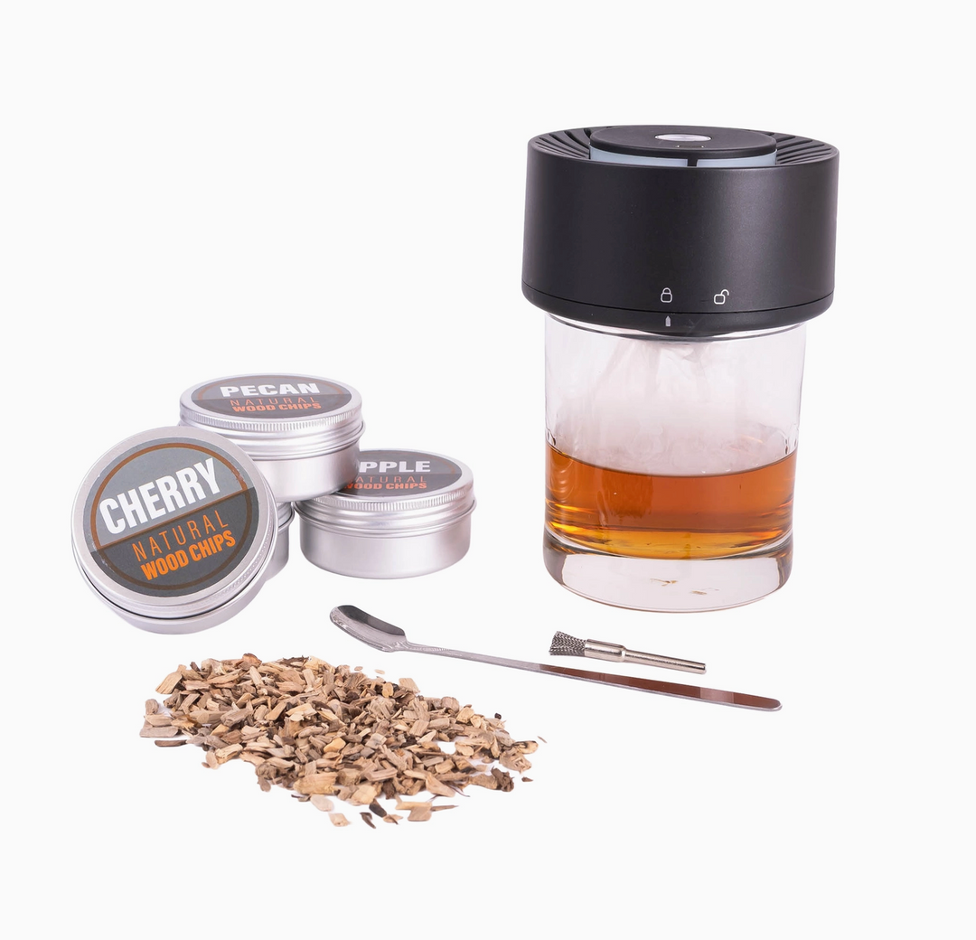 A portable glass vaporizer with a black top and clear lower chamber filled with amber liquid, shown with three silver tin canisters labeled Cherry, Pecan, and Apple wood chips, a metal spoon, a metal tool, and scattered dried wood chips on a white background.