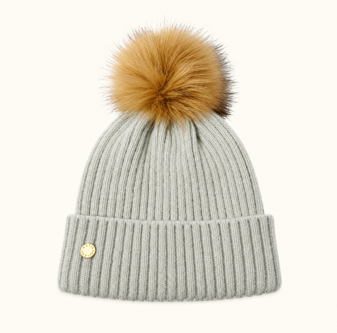 Katie Loxton Knitted Hat in Cool Grey with ribbed knit design, folded cuff, gold logo charm, and tan faux fur pom pom.