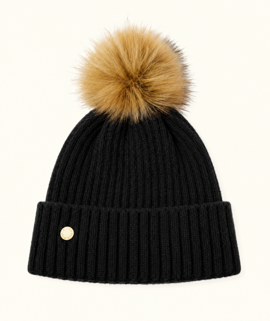 Katie Loxton Knitted Hat in Black with ribbed knit design, folded cuff, gold logo charm, and tan faux fur pom pom.